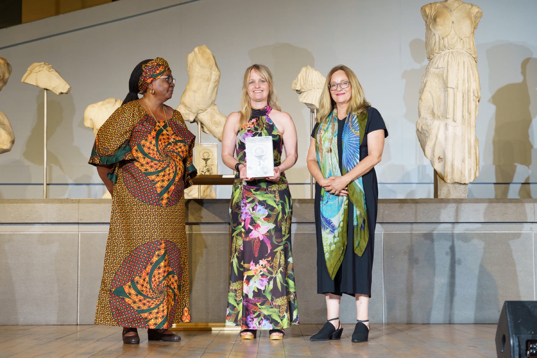 Three women on stage, the laureate at the centre holding her prize