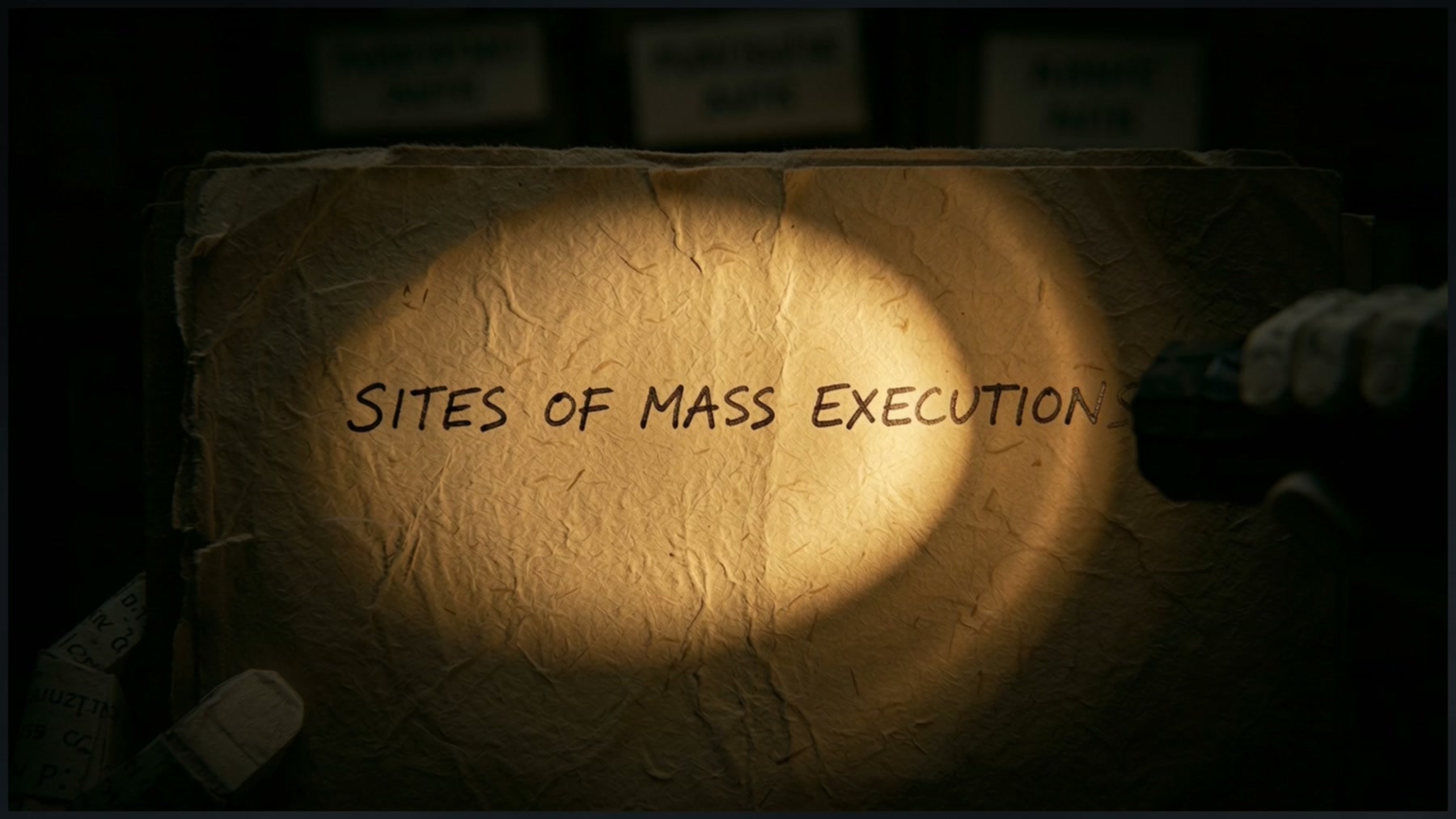 Film still: a flashlight beam falling on a folder handwritten Sites of Mass Executions